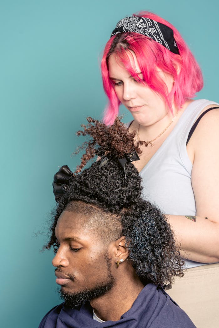 Serious female hairdresser dying hair of African American male sitting in cape against blue background