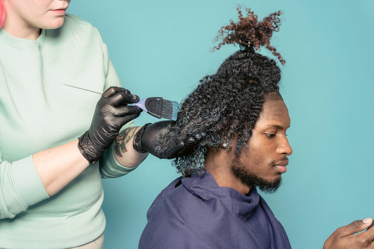about-bg Crop hairstylist with tattoo dying African American male customer using cellphone against blue background